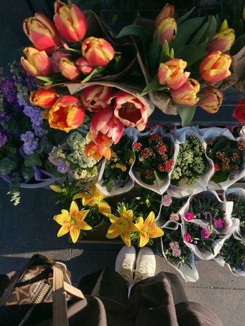 A vibrant display of fresh flower bouquets arranged outdoors, featuring predominantly orange and yellow tulips with ruffled petals and green stems, alongside smaller purple, white, and pink flowers in