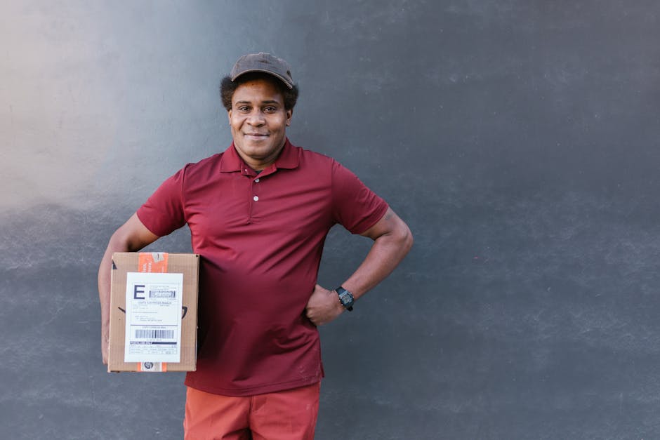 A smiling male florist standing in front of a plain dark grey wall holds a small, rectangular cardboard box with a shipping label, dressed in a maroon polo shirt, matching cap, and red trousers. The f