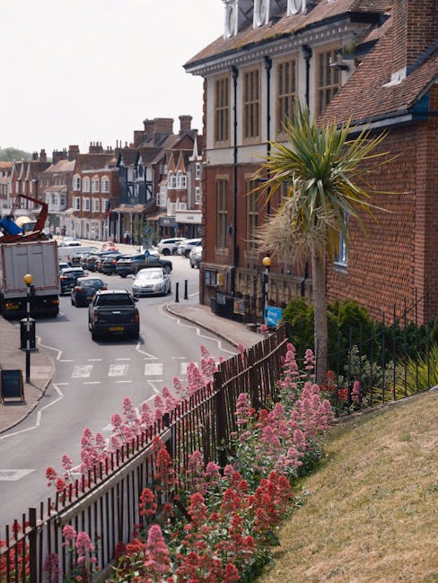 A street scene featuring a Victorian-style building with large bay windows and red brick facade, set against a partly cloudy sky. In the foreground, pink and red flowering plants grow along a black wr